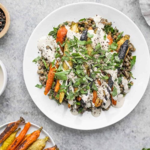A White Plate Filled With Roasted Carrots With Lentils And Fresh Greens Drizzled With Creamy Sauce. On The Side, A Small Bowl Of Peppercorns, A Small Bowl Of Dipping Sauce, And A Serving Dish Boasting Extra Roasted Carrots Complete The Spread.