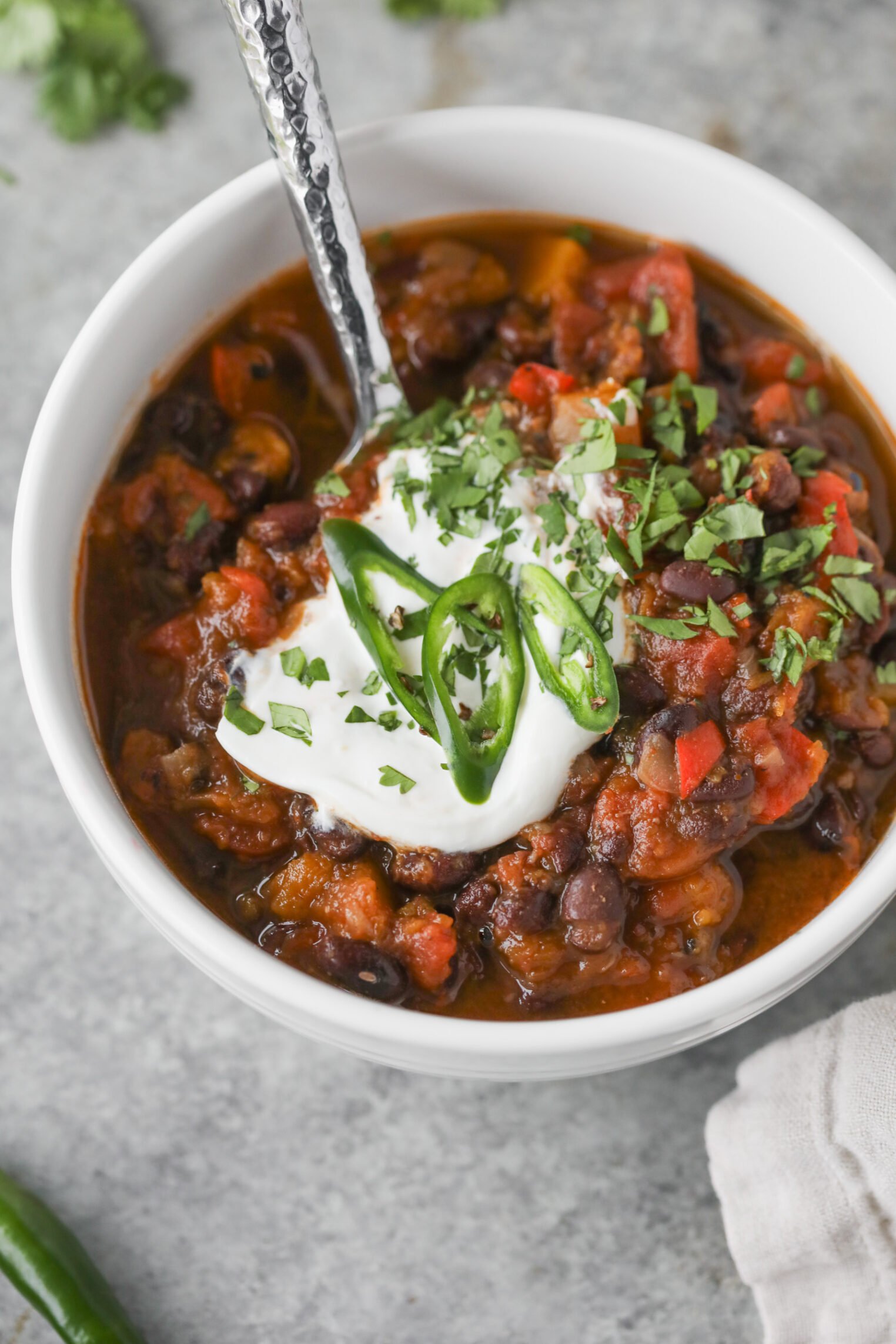 Slow Cooker Black Bean Chili with Butternut Squash 1 A Bowl Of Slow Cooker Vegan Black Bean Chili Topped With A Dollop Of Sour Cream, Sliced Green Chili Peppers, And Chopped Cilantro, With A Spoon Inside. The Chili Contains Beans, Tomatoes, And Visible Vegetables.