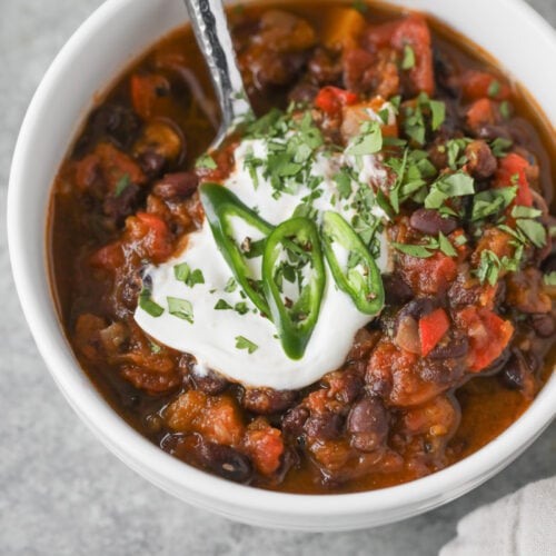 Slow Cooker Black Bean Chili with Butternut Squash 9 A Bowl Of Slow Cooker Vegan Black Bean Chili Topped With A Dollop Of Sour Cream, Sliced Green Chili Peppers, And Chopped Cilantro, With A Spoon Inside. The Chili Contains Beans, Tomatoes, And Visible Vegetables.