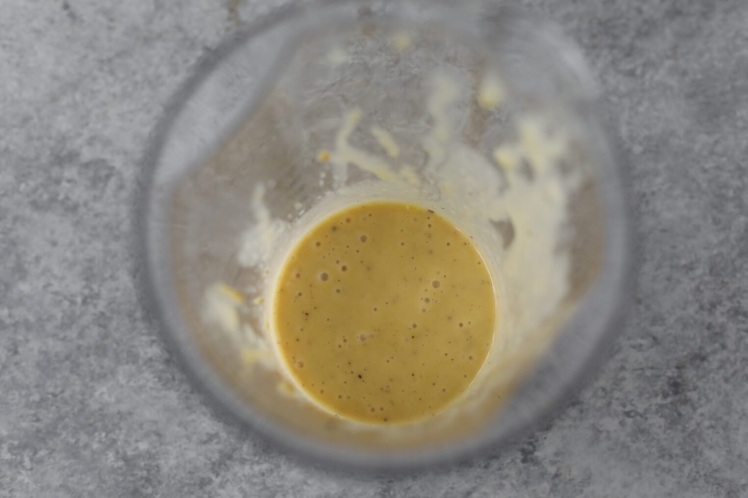 Overhead View Of A Glass Container Holding A Small Amount Of Blended Yellow-Orange Liquid With Bubbles, Likely A Sauce Or Dressing For Wild Rice And Kale Salad, On A Gray Speckled Surface.