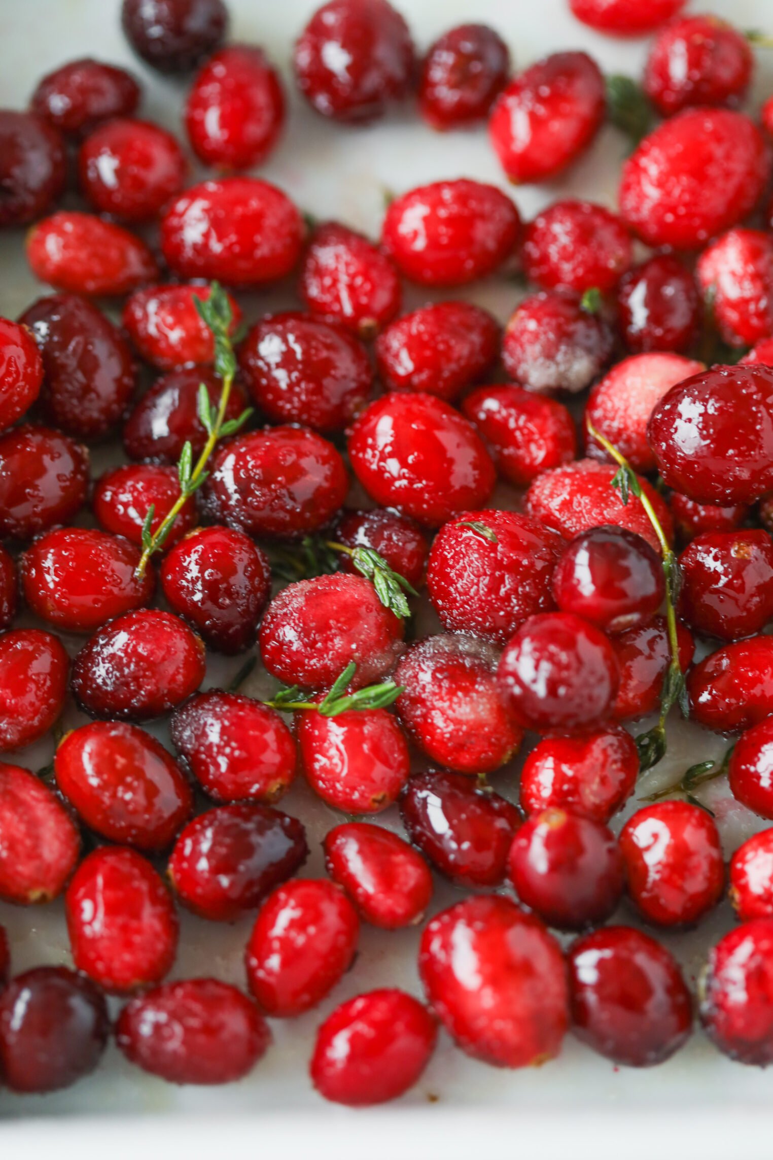 Bright Red Cranberries Coated With A Shiny Glaze Are Scattered On A White Surface, Accented By Sprigs Of Fresh Green Herbs—Perfect As A Garnish Or Vibrant Ingredient For A Wild Rice And Kale Salad.