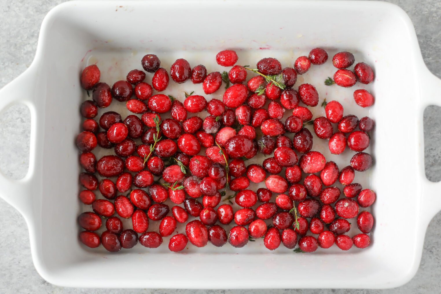 A White Rectangular Baking Dish Filled With Fresh Cranberries Coated In Sugar, Garnished With A Few Sprigs Of Fresh Rosemary, Sits On A Light Gray Countertop—Perfect Alongside A Wild Rice And Kale Salad.