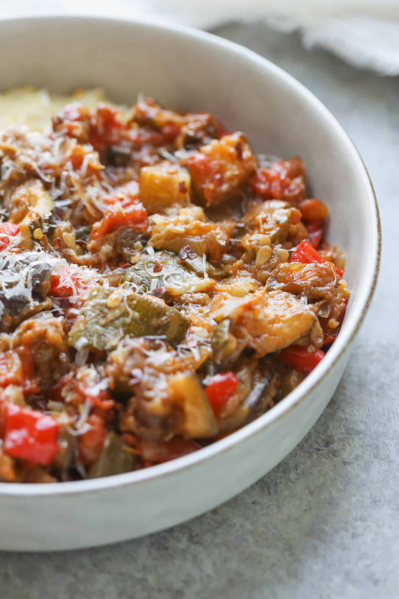 A Close-Up Of A Bowl Filled With Chunky Roasted Ratatouille, Featuring Pieces Of Zucchini, Eggplant, Tomatoes, And Red Bell Peppers, All Topped With Grated Cheese.