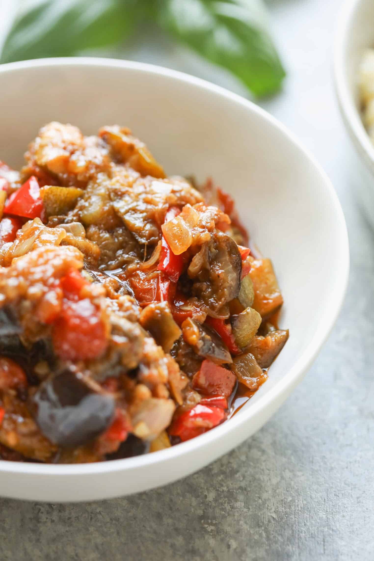 A White Bowl Filled With Roasted Ratatouille, A Vegetable Stew Featuring Chunks Of Eggplant, Red Bell Pepper, Tomatoes, And Onions In A Rich Sauce, With Fresh Basil Leaves Blurred In The Background.