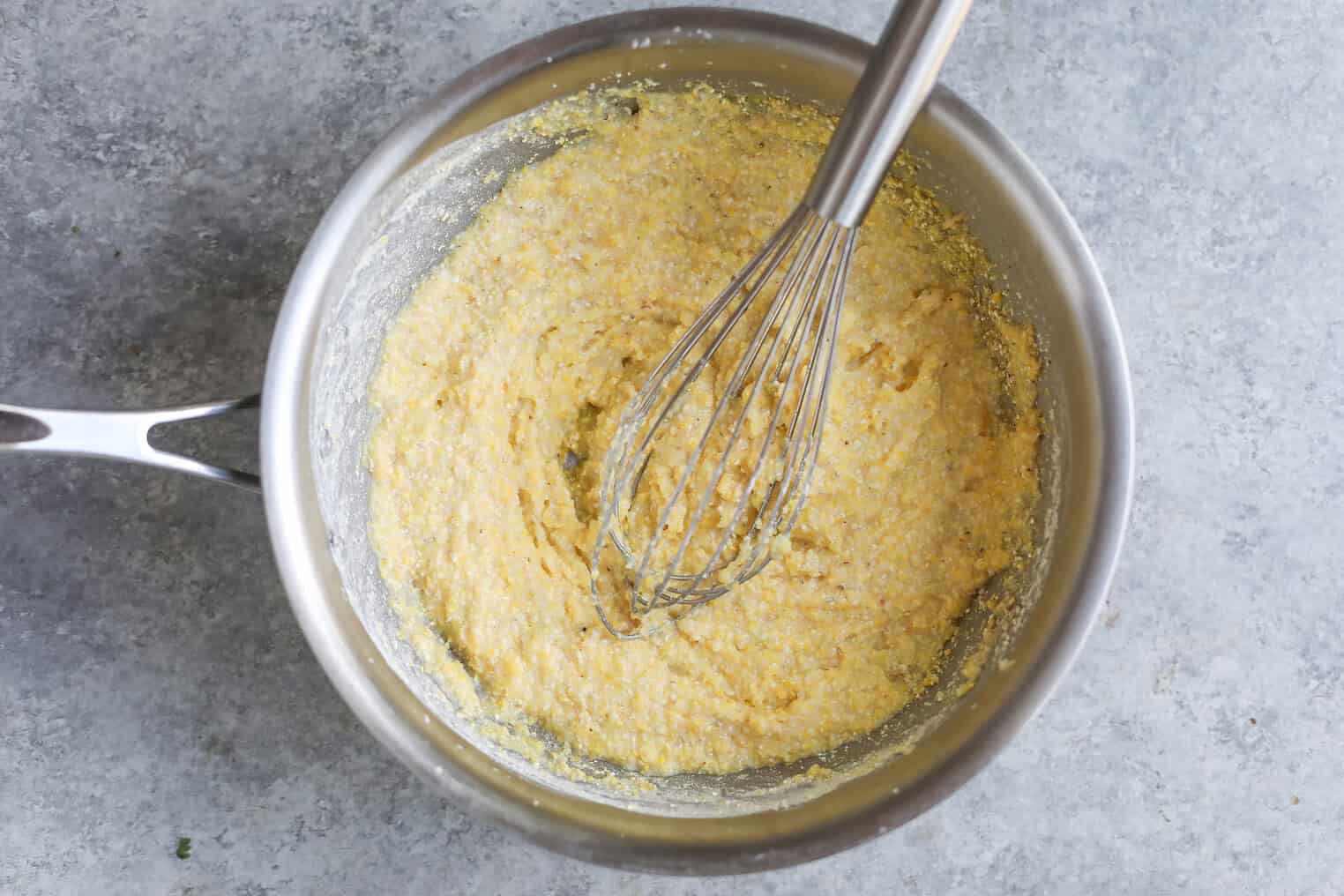 A Metal Mixing Bowl Filled With A Thick, Yellow Batter Being Mixed With A Metal Whisk, Set On A Gray Countertop, Ready For Pairing With Roasted Ratatouille.