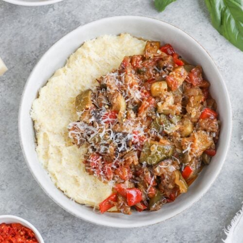A Bowl Of Creamy Polenta Topped With A Colorful Roasted Ratatouille Of Diced Vegetables And Grated Cheese, Garnished With Fresh Basil, With Small Dishes Of Ingredients Nearby On A Light Gray Surface.
