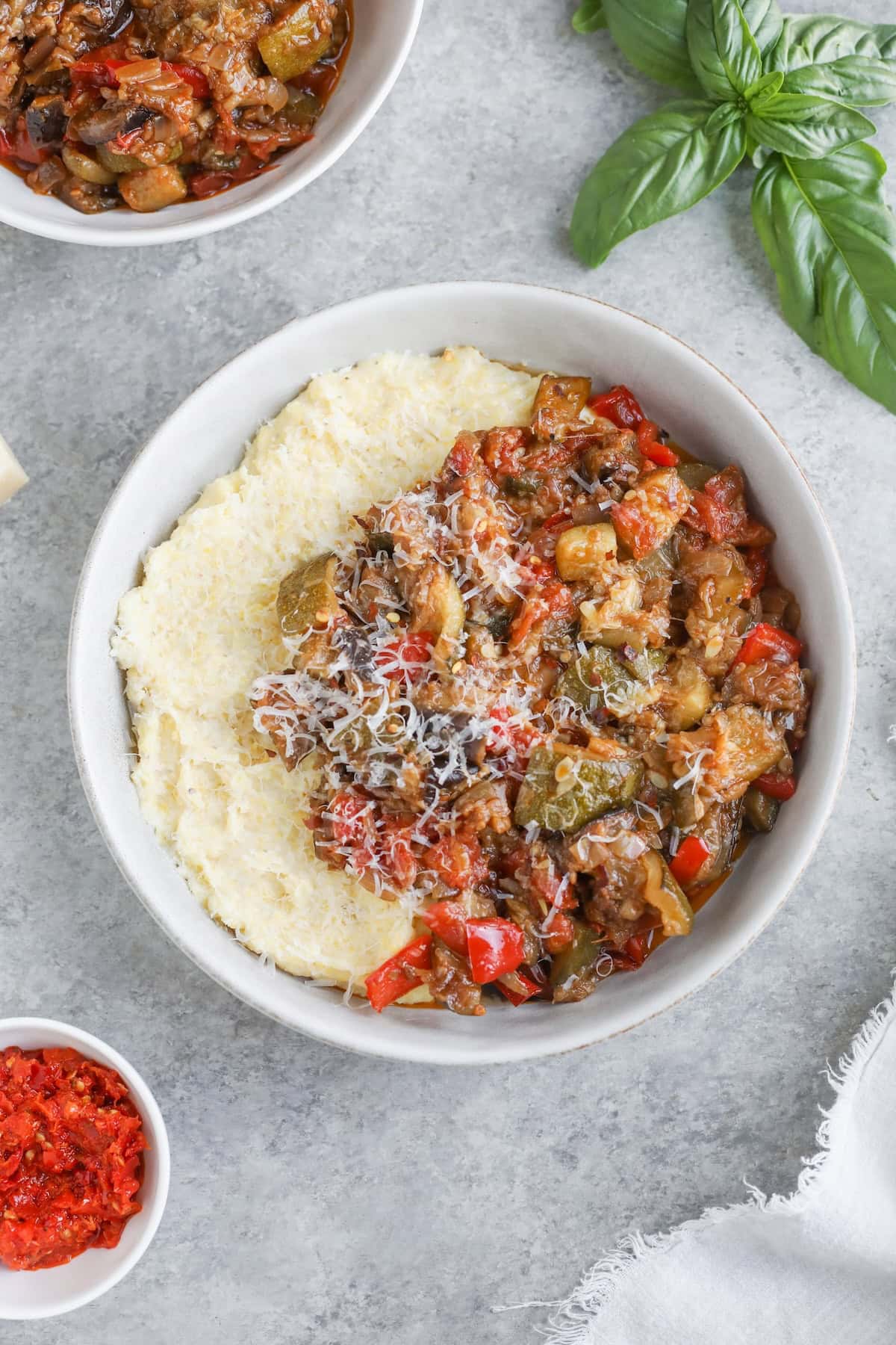 A Bowl Of Creamy Polenta Topped With A Colorful Roasted Ratatouille Of Diced Vegetables And Grated Cheese, Garnished With Fresh Basil, With Small Dishes Of Ingredients Nearby On A Light Gray Surface.