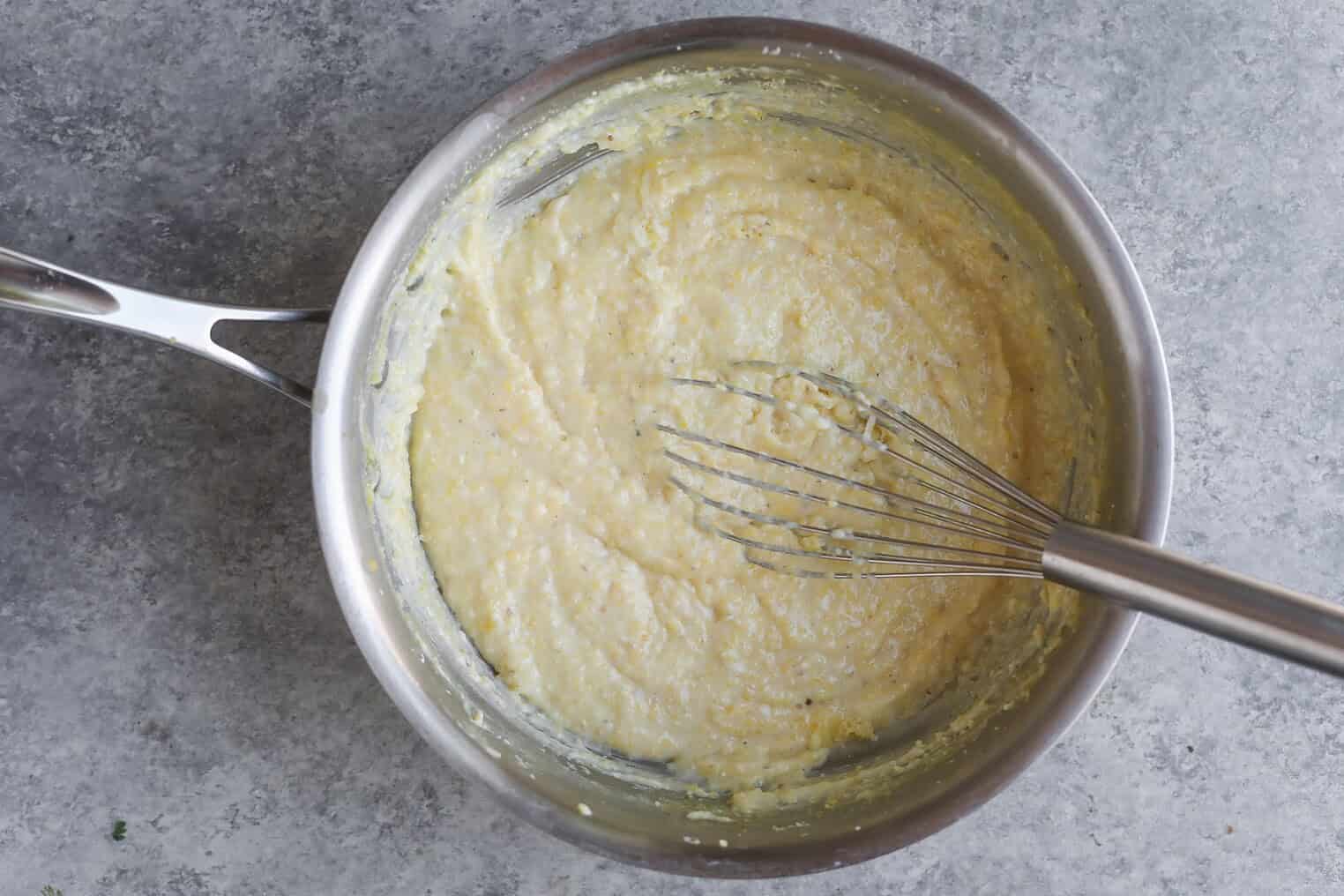 A Metal Saucepan Containing Creamy, Yellow Grits Being Mixed With A Metal Whisk, Set On A Gray Textured Surface, Beside A Bowl Of Vibrant Roasted Ratatouille.