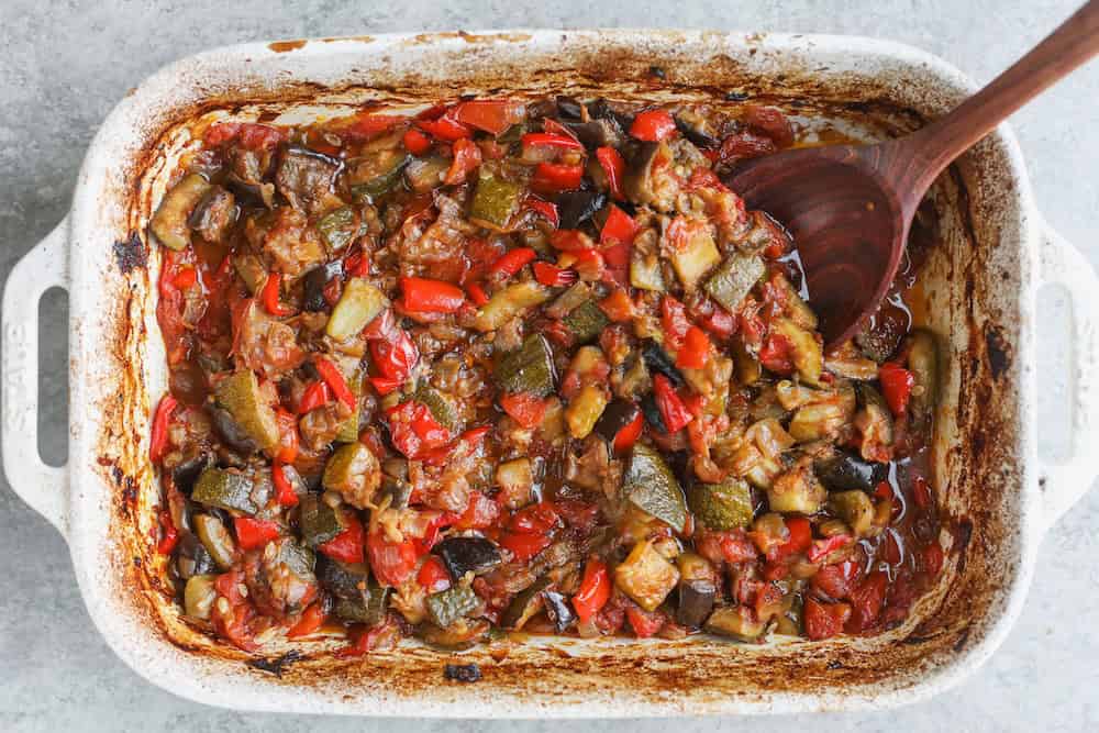 A White Baking Dish Filled With Roasted Ratatouille Featuring Chopped Eggplant, Zucchini, Tomatoes, And Red Bell Peppers, With A Wooden Spoon Resting Inside. The Edges Of The Dish Are Perfectly Browned From Baking.