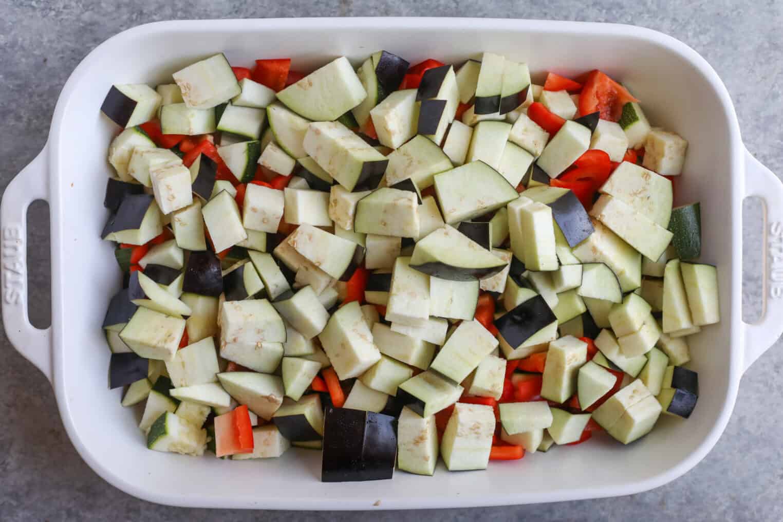 A White Baking Dish Filled With Chopped Raw Vegetables, Including Eggplant, Zucchini, And Red Bell Pepper, Sits On A Gray Surface—Perfect For Making Roasted Ratatouille.