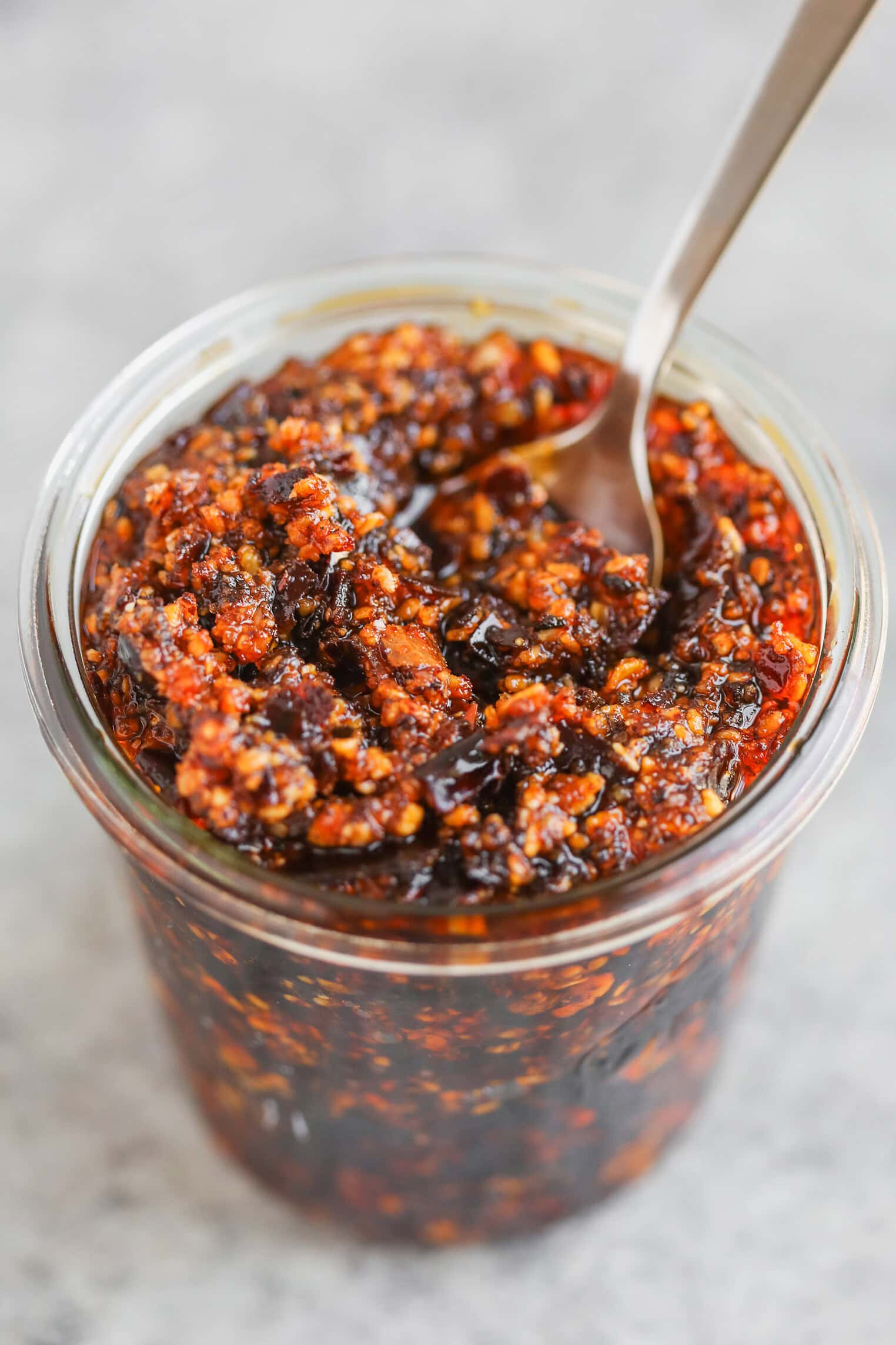 A Glass Jar Filled With Chunky Chili Oil, Reminiscent Of Salsa Macha, Containing Red Pepper Flakes And Bits Of Garlic, With A Metal Spoon Inside. The Background Is Out Of Focus.
