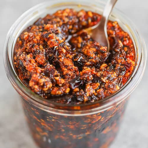 A Glass Jar Filled With Chunky Chili Oil, Reminiscent Of Salsa Macha, Containing Red Pepper Flakes And Bits Of Garlic, With A Metal Spoon Inside. The Background Is Out Of Focus.