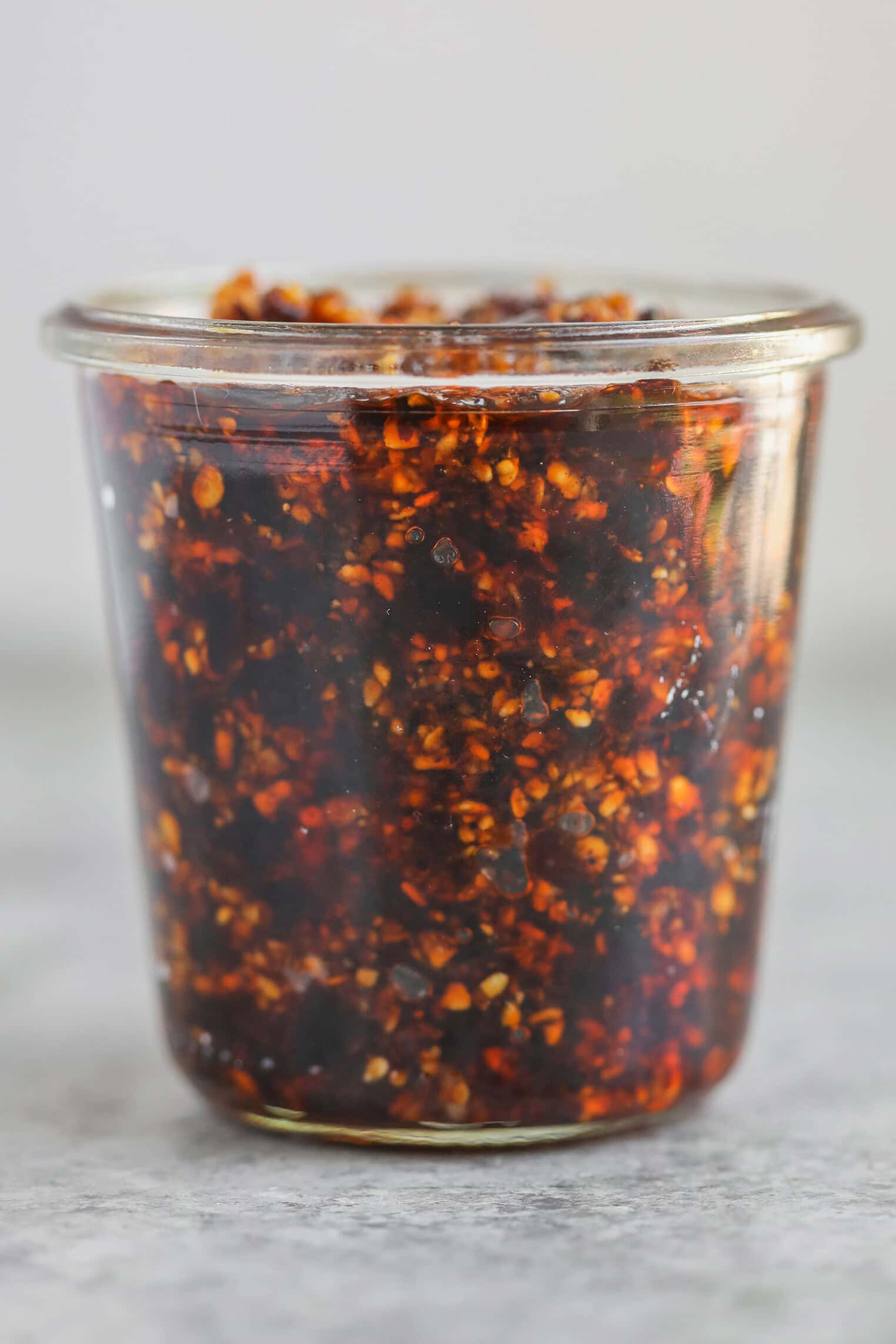 A Clear Glass Jar Filled With Chunky Salsa Macha, Showing Visible Chili Flakes And Seeds In A Rich, Red-Brown Oil. The Jar Is Placed On A Light Gray Surface With A Neutral Background.
