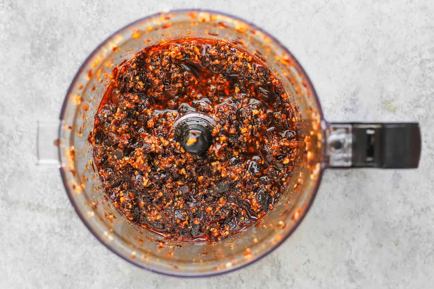Overhead View Of A Food Processor Containing A Coarse, Red Chili Paste Resembling Salsa Macha, With Visible Pepper Seeds And Oil On A Light Gray Surface.