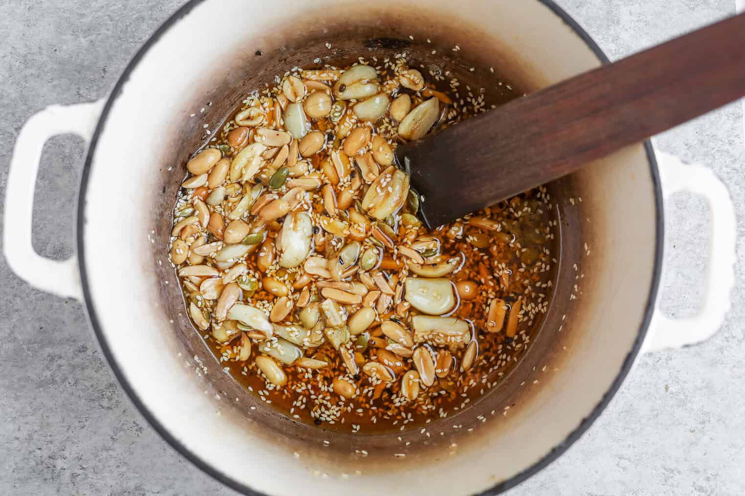 A White Pot Containing Garlic Cloves, Peanuts, Sesame Seeds, And Oil Being Stirred With A Dark Wooden Spatula, Viewed From Above On A Light Gray Surface—A Classic Start To Homemade Salsa Macha.