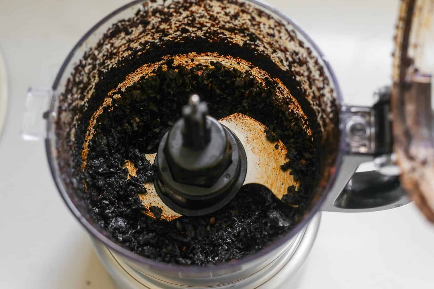 A Close-Up Of A Food Processor With Dark, Burnt Salsa Macha Residue And Chunks Stuck To The Sides And Blade, Indicating It Has Been Overprocessed Or Burned.