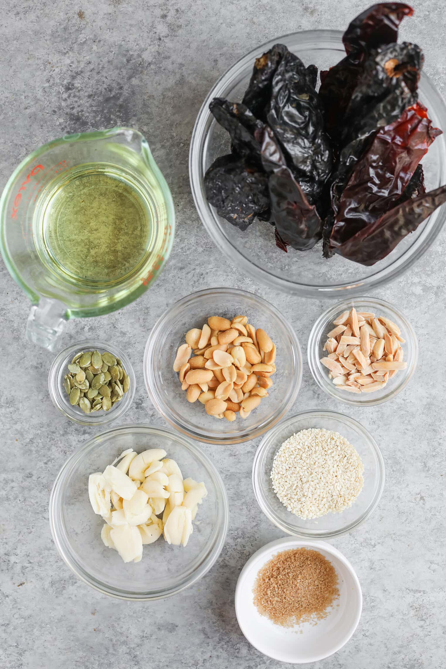 A Variety Of Ingredients For Salsa Macha In Small Glass Bowls On A Gray Surface, Including Dried Chilies, Oil, Pepitas, Peanuts, Almonds, Sesame Seeds, Garlic Cloves, And Brown Sugar.