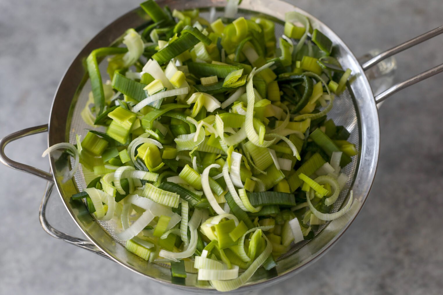 Chopped Leeks In A Metal Strainer, With Green And White Pieces Visible, Set On A Gray Countertop—Perfect For Adding To Savory Oatmeal.