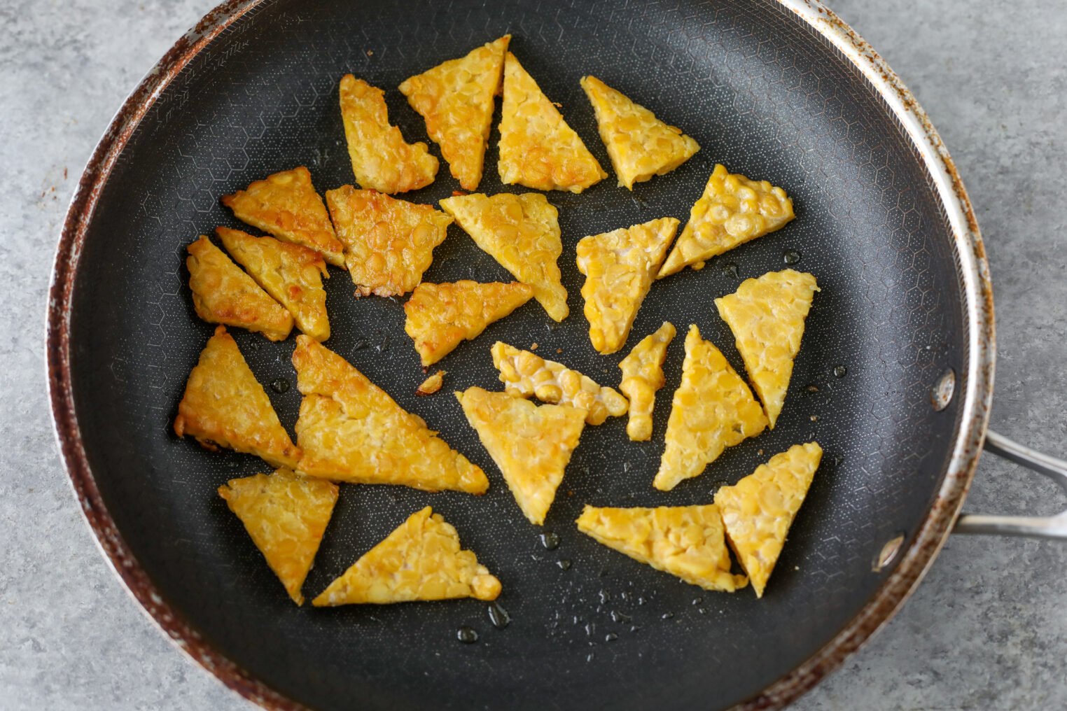 Sliced Triangular Pieces Of Tempeh Are Being Pan-Fried In A Nonstick Skillet, With Some Pieces Golden Brown And Others Slightly Lighter In Color.