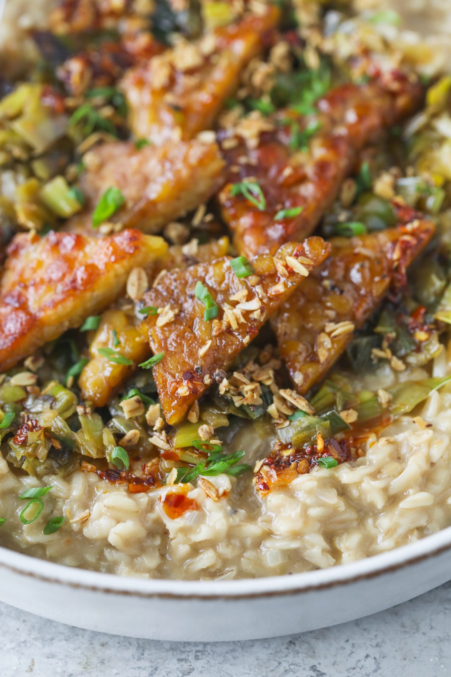A Close-Up Of A Bowl Filled With Creamy Oatmeal Topped With Crispy, Glazed Tofu Slices, Chopped Green Onions, And Bits Of Cooked Vegetables.
