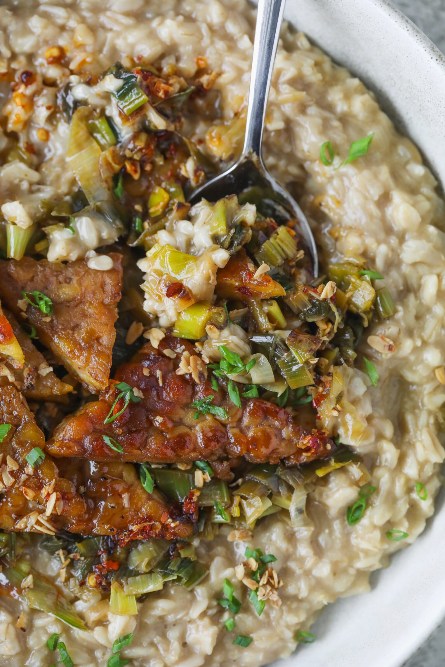 A Close-Up Of A Creamy Risotto Topped With Glazed Tempeh, Sautéed Leeks, And Chopped Green Herbs, With A Spoon Scooping Up A Portion.