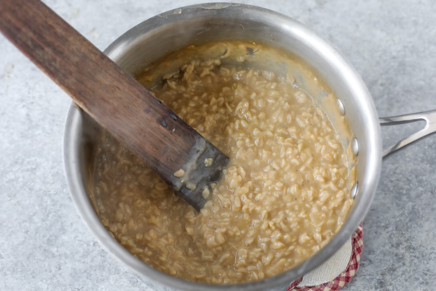 A Pot Of Creamy Cooked Oatmeal Being Stirred With A Wooden Spatula On A Light Gray Surface.