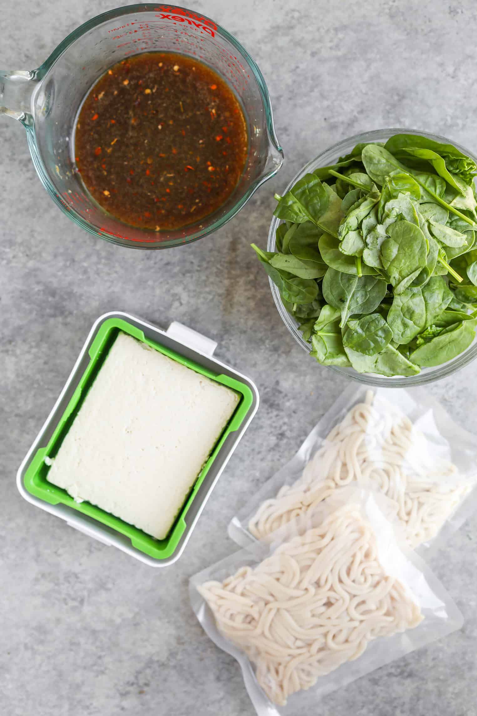 Sesame Garlic Stir Fry Noodles 2 A Top-Down View Of Stir Fry Noodles With A Measuring Cup Of Dark Sauce, A Bowl Of Fresh Spinach, A Block Of Tofu In A Container, And Two Packets Of Uncooked Noodles On A Gray Surface.