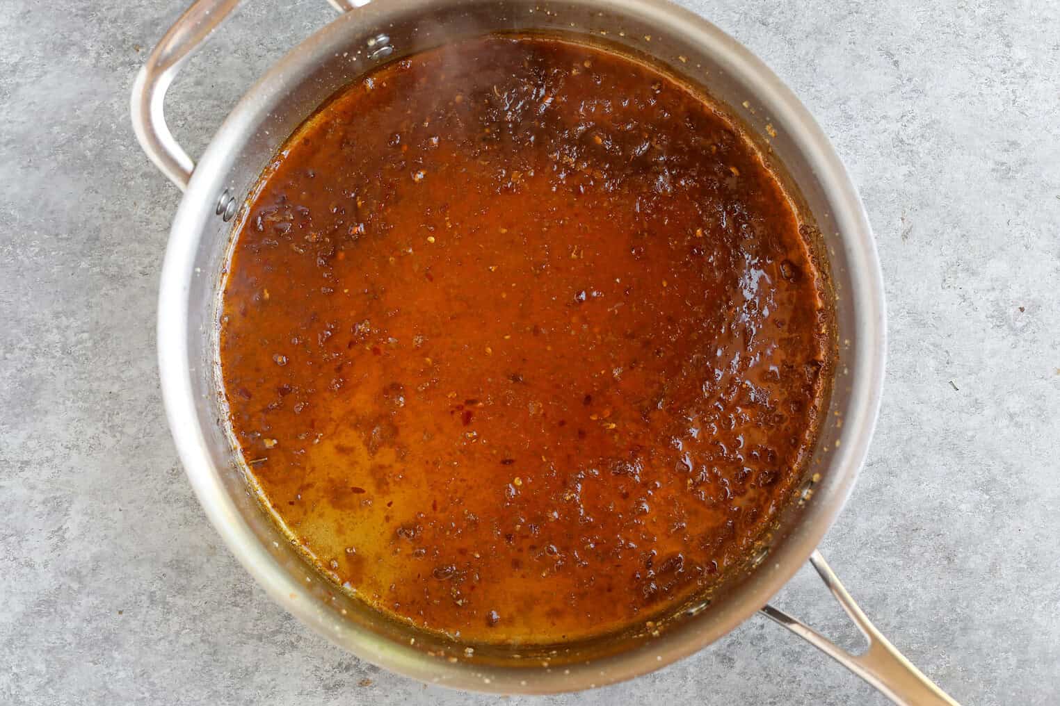 Sesame Garlic Stir Fry Noodles 8 A Stainless Steel Saucepan Filled With Reddish-Brown Simmering Sauce, Likely A Reduction For Stir Fry Noodles With Visible Seasoning Particles, Sits On A Gray Countertop.