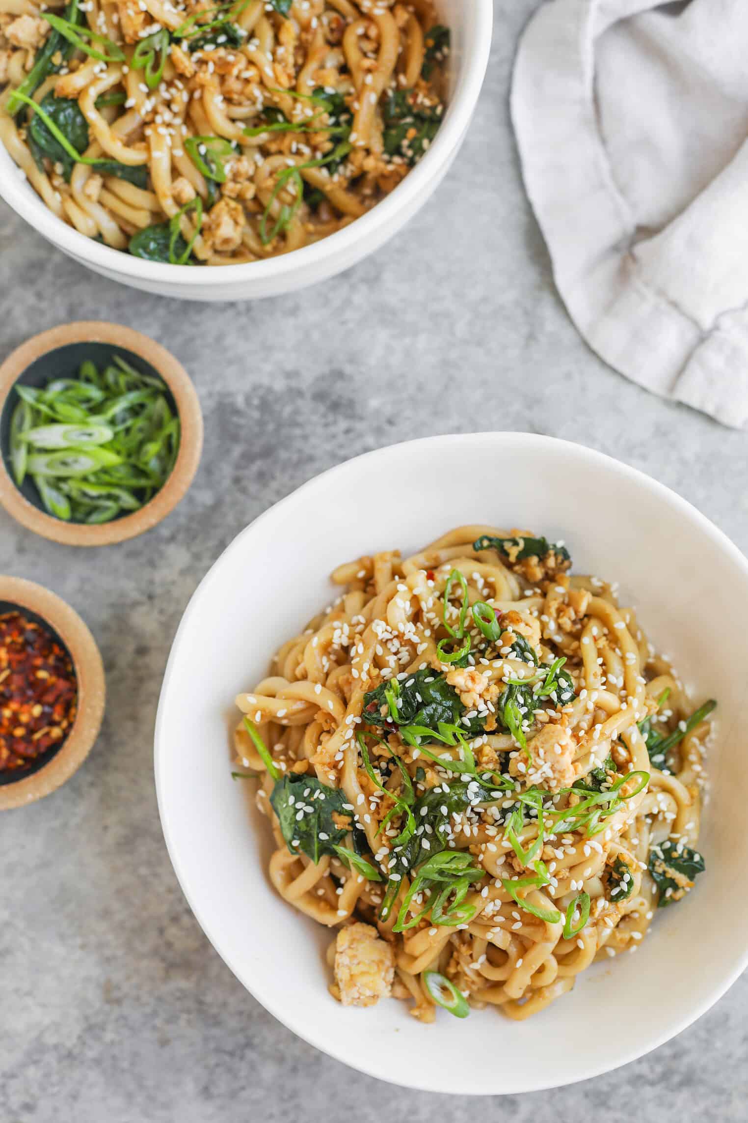 Sesame Garlic Stir Fry Noodles 11 Two White Bowls Filled With Stir Fry Noodles, Greens, And Sesame Seeds Sit On A Gray Surface. Small Bowls With Sliced Green Onions And Chili Flakes Are Nearby, Along With A White Cloth Napkin.