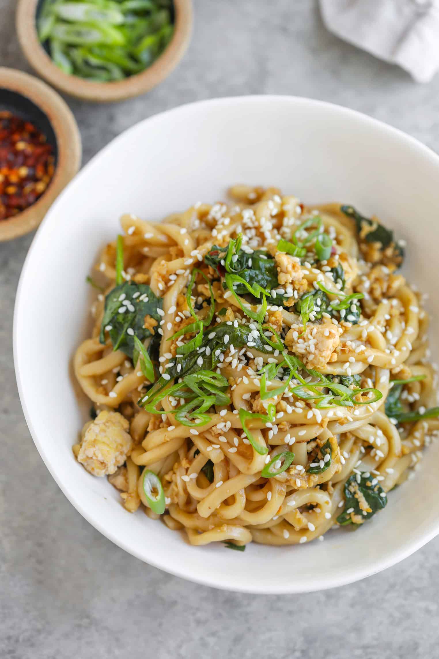 A Bowl Of Udon Stir Fry Noodles With Greens, Topped With Sesame Seeds And Sliced Green Onions. Small Bowls Of Chili Flakes And Extra Green Onions Are Nearby On A Light, Textured Surface.