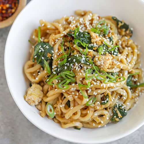 Sesame Garlic Stir Fry Noodles 13 A Bowl Of Udon Stir Fry Noodles With Greens, Topped With Sesame Seeds And Sliced Green Onions. Small Bowls Of Chili Flakes And Extra Green Onions Are Nearby On A Light, Textured Surface.