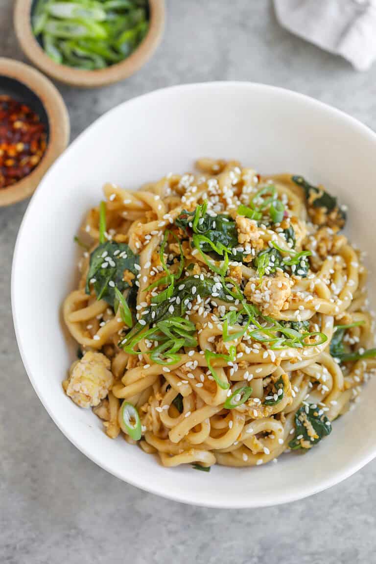 A Bowl Of Udon Stir Fry Noodles With Greens, Topped With Sesame Seeds And Sliced Green Onions. Small Bowls Of Chili Flakes And Extra Green Onions Are Nearby On A Light, Textured Surface.