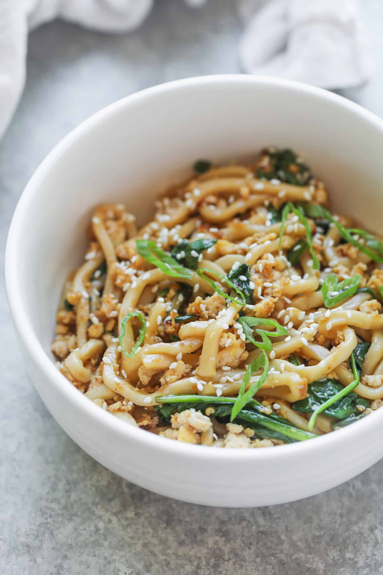 Sesame Garlic Stir Fry Noodles 10 A White Bowl Filled With Stir Fry Noodles, Topped With Green Onions, Spinach, And Sesame Seeds, Sits On A Gray Surface.