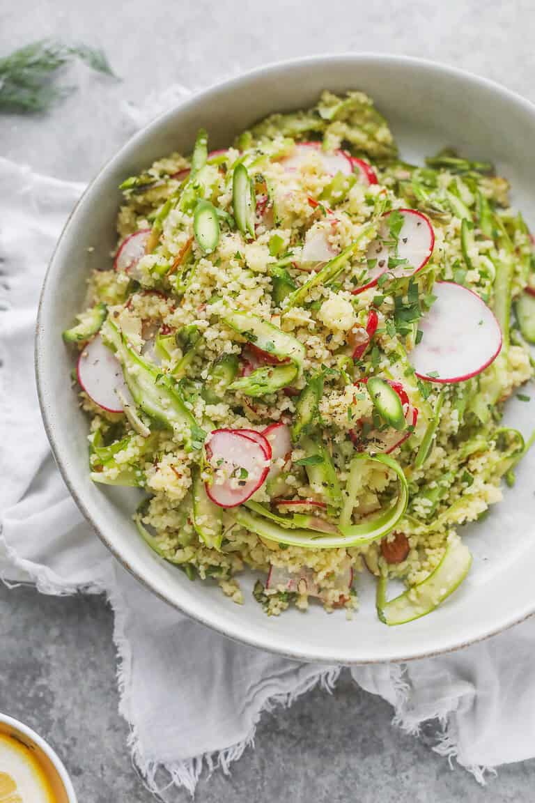 A Bowl Of Couscous Features A Vibrant Shaved Asparagus Spring Salad, With Thinly Sliced Radishes And Fresh Herbs, Served On A Light Gray Surface With A White Cloth Underneath.