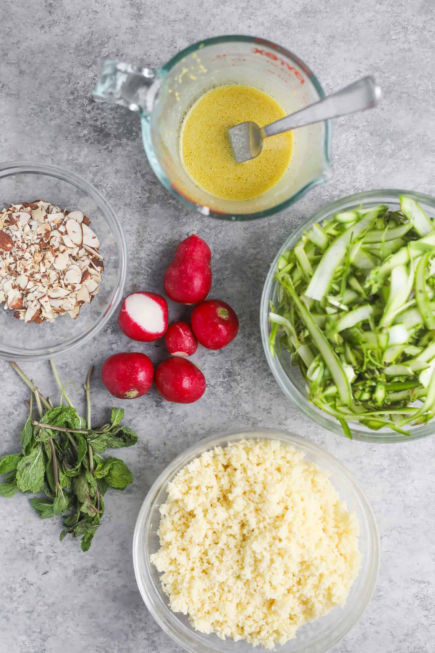 Shaved Asparagus Spring Salad 3 Ingredients For A Shaved Asparagus Spring Salad On A Gray Surface: Sliced Almonds, Fresh Mint, Whole And Halved Radishes, Shredded Asparagus, Cooked Couscous, And A Glass Measuring Cup With Dressing And A Spoon.