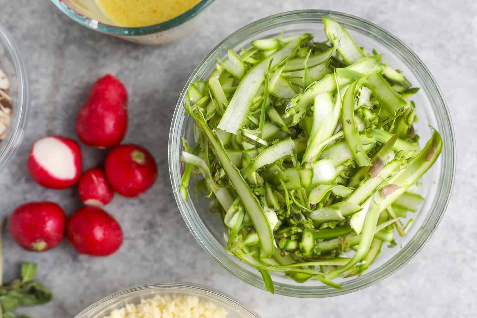 Shaved Asparagus Spring Salad 4 A Glass Bowl Filled With Shaved Asparagus Spring Salad Sits On A Gray Surface Next To Whole Red Radishes And Parts Of Other Ingredient Bowls.