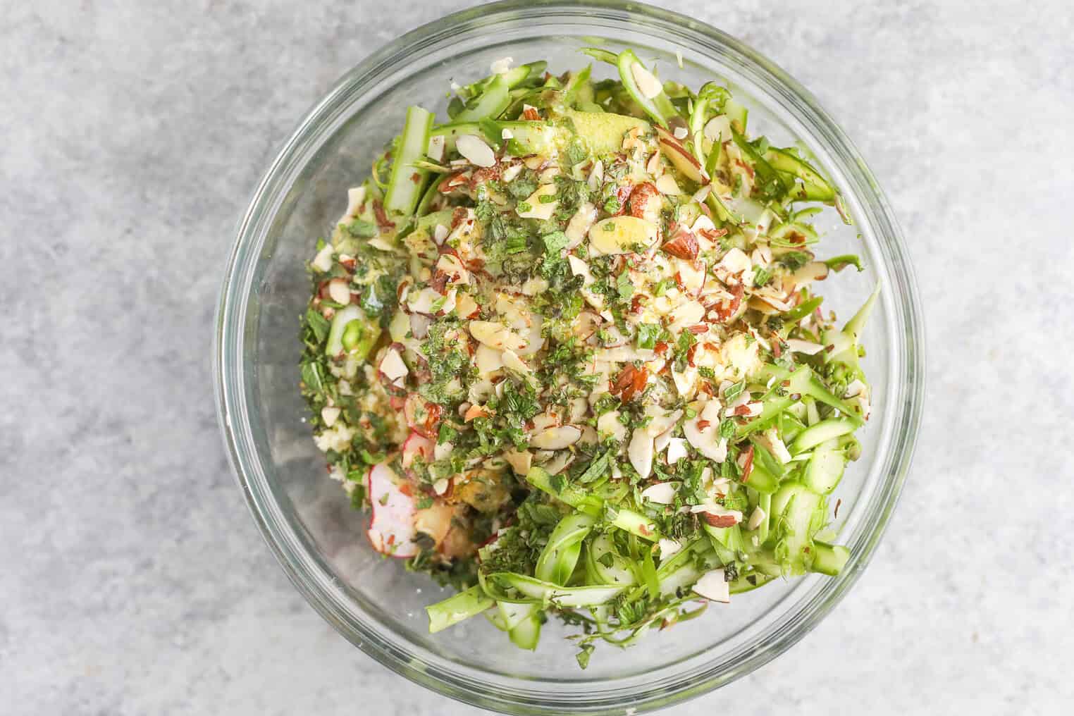 Shaved Asparagus Spring Salad 7 A Glass Bowl Filled With A Shaved Asparagus Spring Salad, Featuring Mixed Greens Topped With Sliced Almonds And Chopped Herbs, Set On A Light Gray Surface.