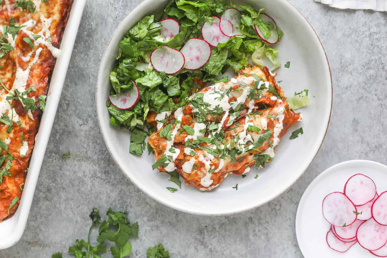 Bean and Cheese Enchiladas 2 A Plate With Two Bean And Cheese Enchiladas Topped With White Sauce And Chopped Herbs, Served With A Side Of Lettuce And Sliced Radishes. Additional Sliced Radishes Are On A Small Plate Next To The Main Dish.