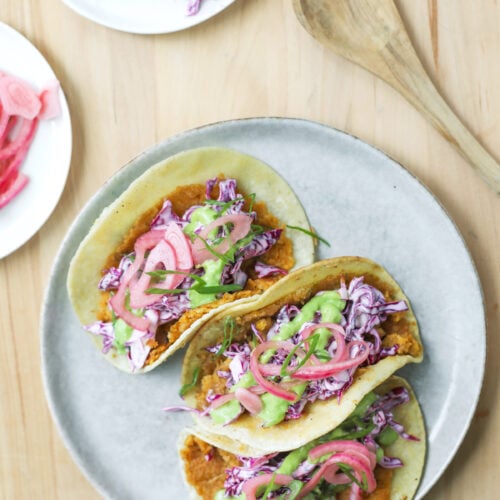 Smashed Chickpea Tacos 12 Three Smashed Chickpea Tacos On A Plate Filled With Fried Toppings, Shredded Purple Cabbage, Pickled Onions, And Green Sauce, Placed On A Wooden Table With A Spoon And Extra Cabbage Nearby.
