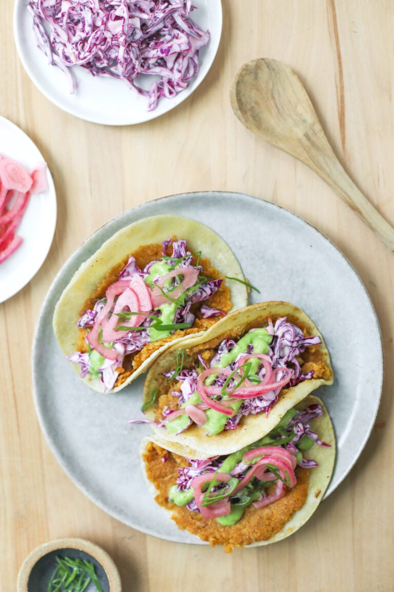 Three Smashed Chickpea Tacos On A Plate Filled With Fried Toppings, Shredded Purple Cabbage, Pickled Onions, And Green Sauce, Placed On A Wooden Table With A Spoon And Extra Cabbage Nearby.
