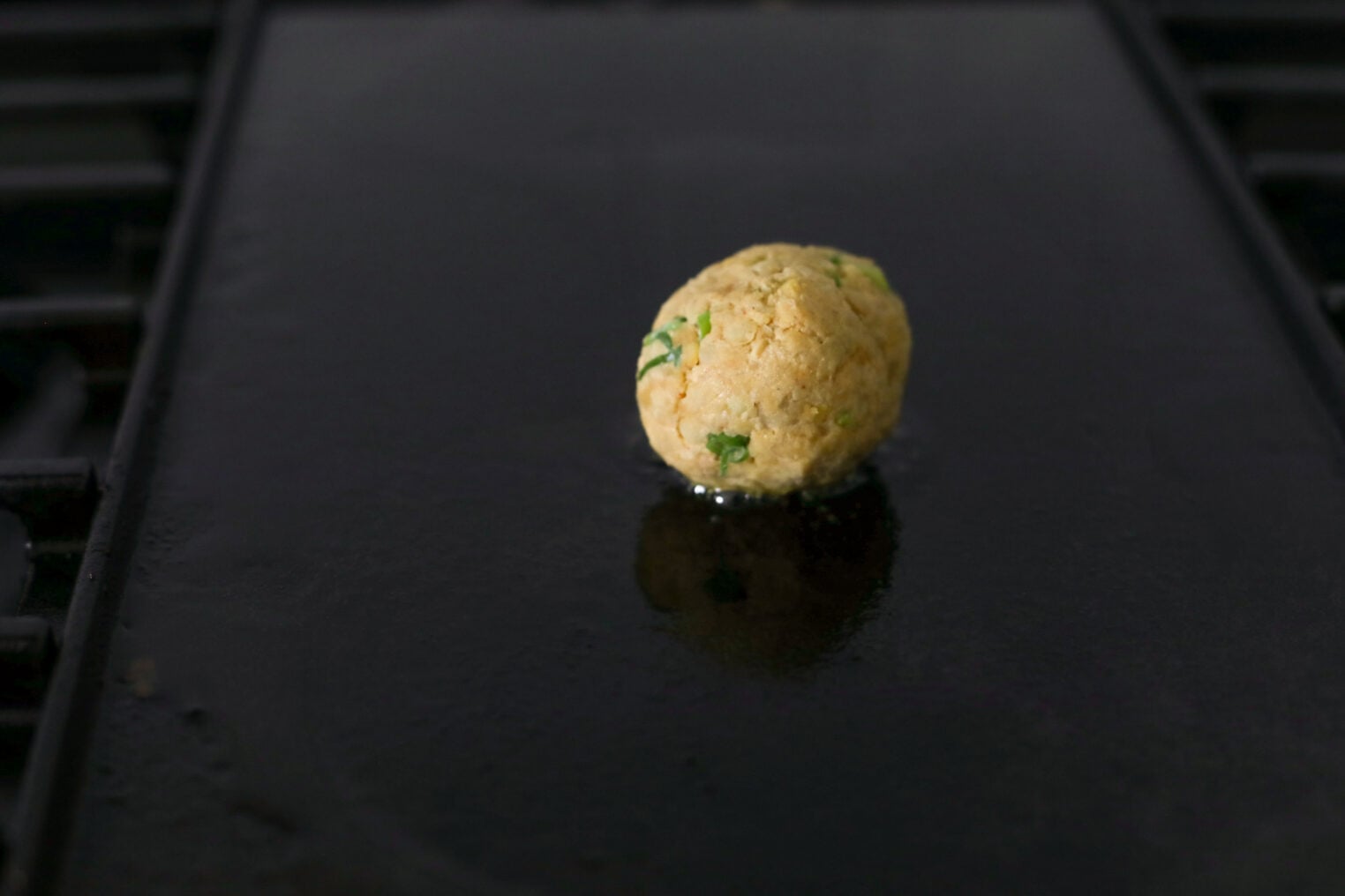 Smashed Chickpea Tacos 7 A Single Uncooked Falafel Ball With Visible Herbs Sits On A Dark, Oiled Griddle, Ready To Be Cooked—A Tempting Sight Reminiscent Of The Flavors Found In Smashed Chickpea Tacos.