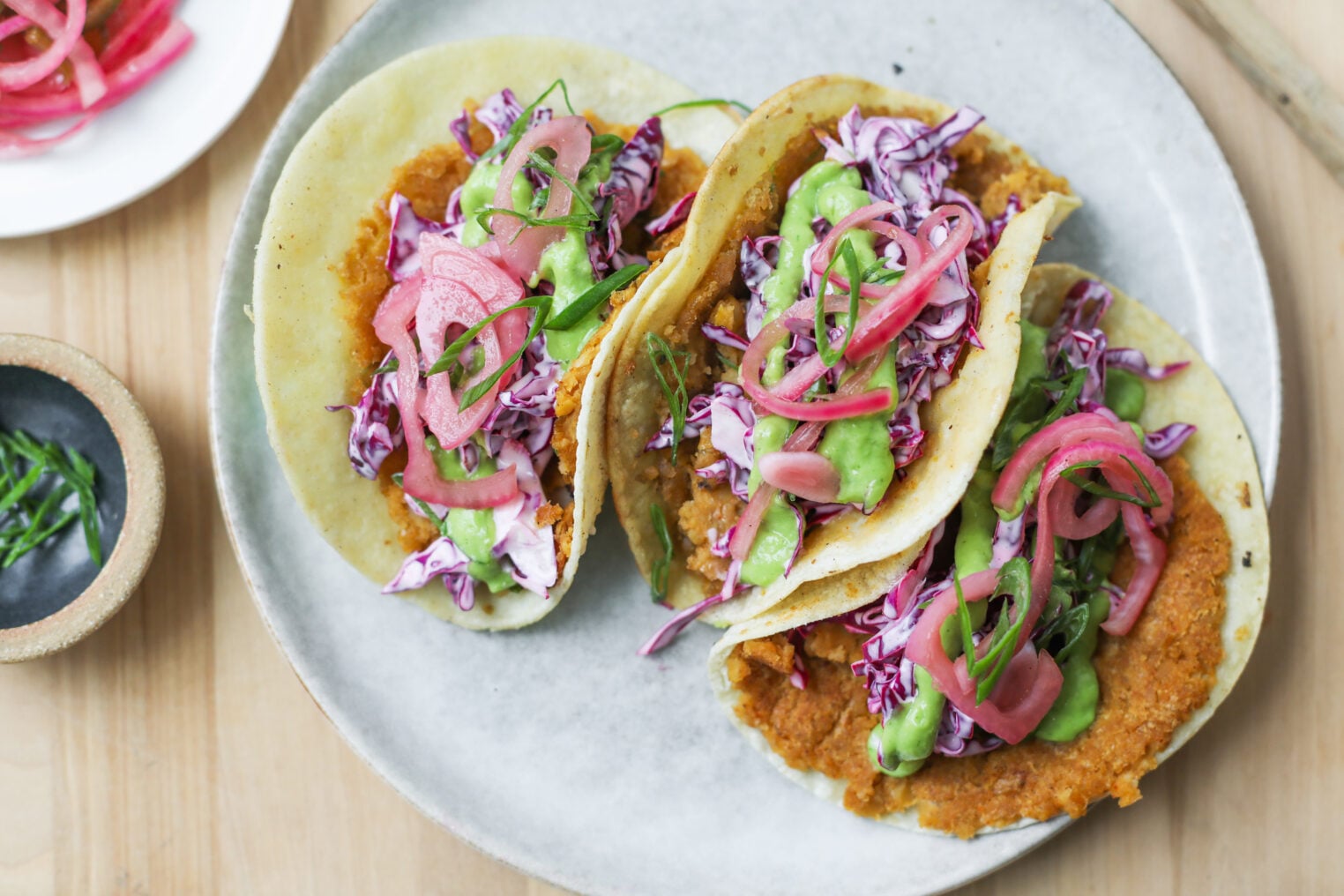 Smashed Chickpea Tacos 9 Three Smashed Chickpea Tacos Filled With Shredded Purple Cabbage, Avocado, And Pickled Red Onions Are Arranged On A White Plate. A Small Bowl With Fresh Herbs Sits Nearby On A Wooden Table.