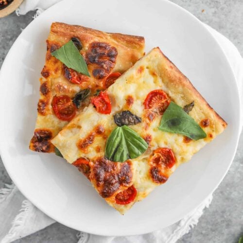 Two Slices Of Margherita Pizza With Melted Cheese, Cherry Tomatoes, And Fresh Basil Rest On A White Plate. The Crust, Made From Sourdough, Adds A Delightful Tang. The Plate Is Set On A Gray Surface With A White Cloth Beside It.