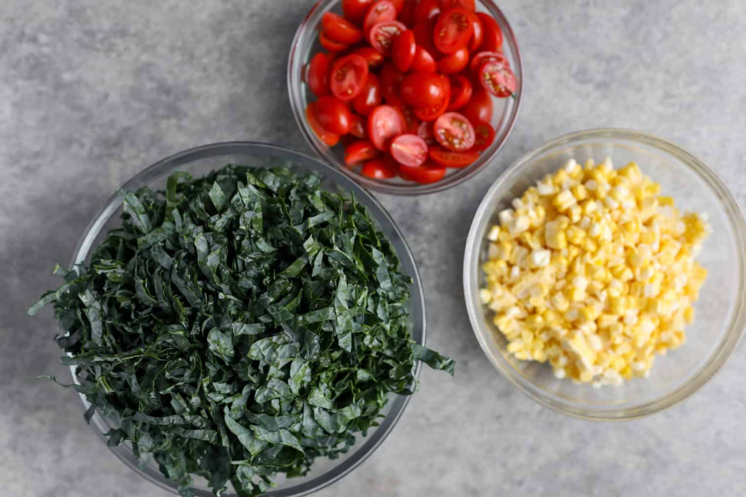 Spicy Kale Salad with Avocado Ranch 4 Three Glass Bowls On A Gray Surface: One With Spicy Kale Salad Made From Sliced Dark Leafy Greens, One With Halved Cherry Tomatoes, And One With Chopped Yellow Corn.