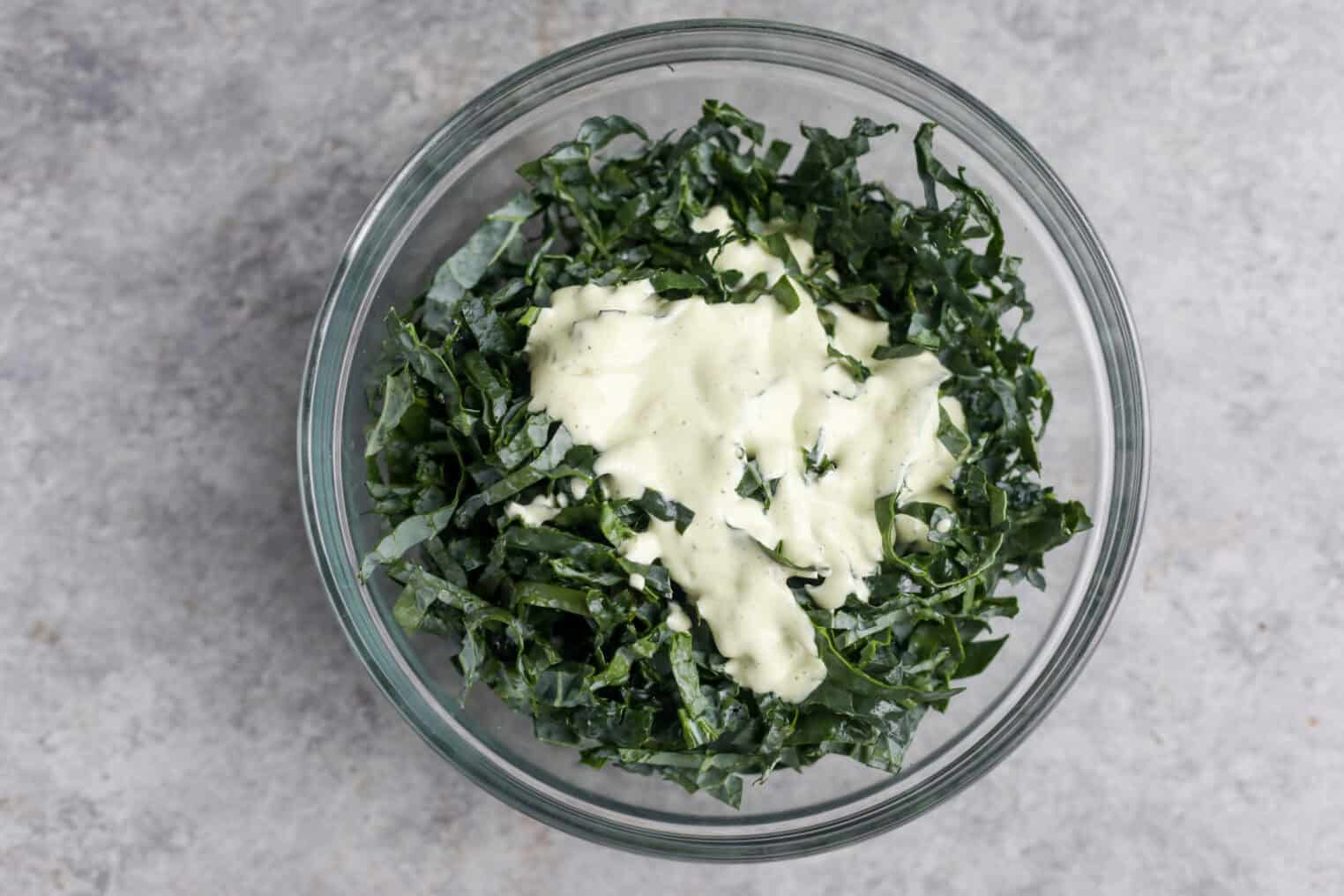 Spicy Kale Salad with Avocado Ranch 6 A Glass Bowl Filled With Chopped Dark Green Spicy Kale Salad, Topped With A Dollop Of Creamy White Dressing, Sits On A Light Gray Textured Surface.