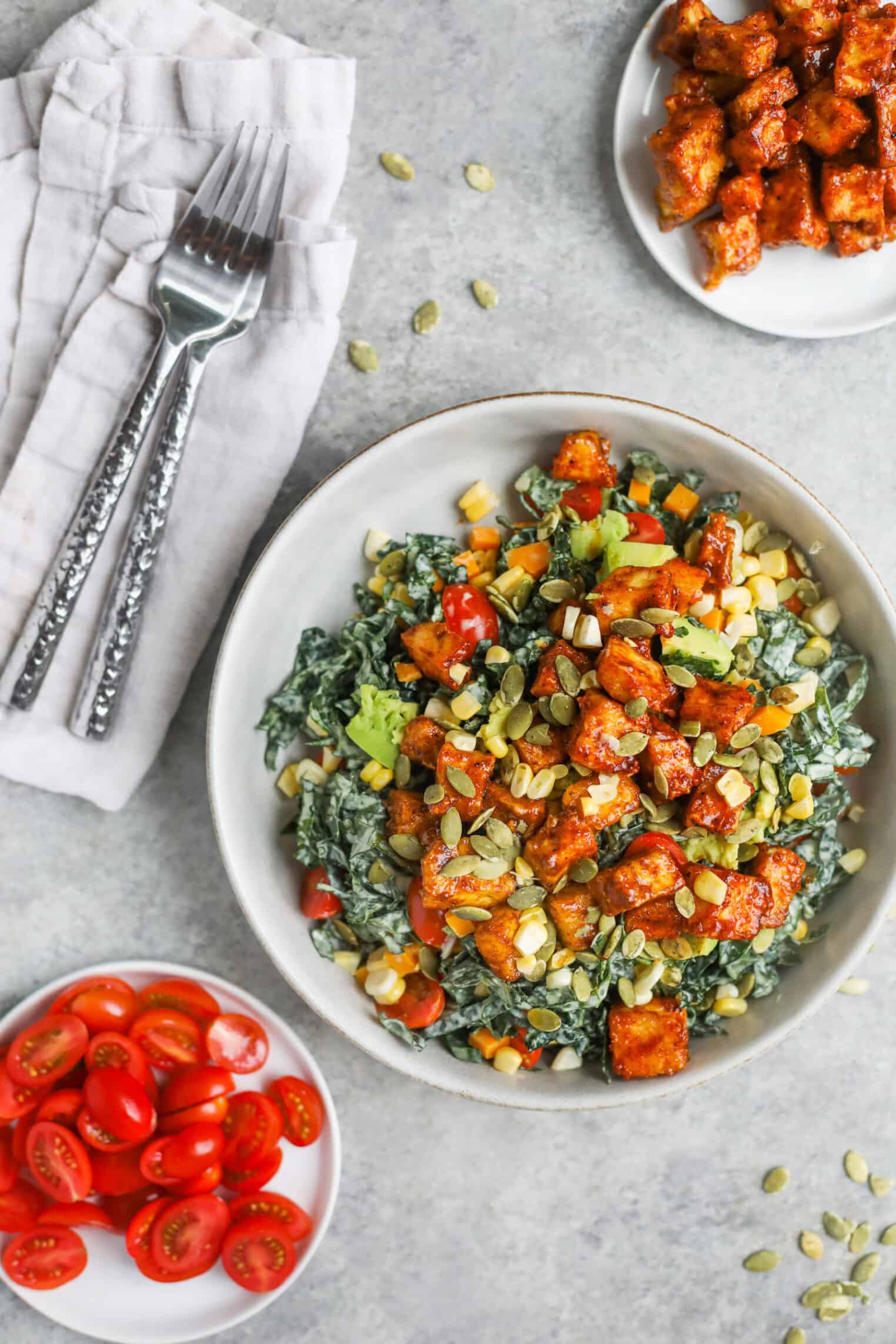 Spicy Kale Salad with Avocado Ranch 2 A Bowl Of Spicy Kale Salad With Cherry Tomatoes, Corn, Seeds, And Roasted Tofu, Surrounded By Small Plates Of Sliced Cherry Tomatoes And Tofu Cubes, With A Napkin And Forks On The Side.