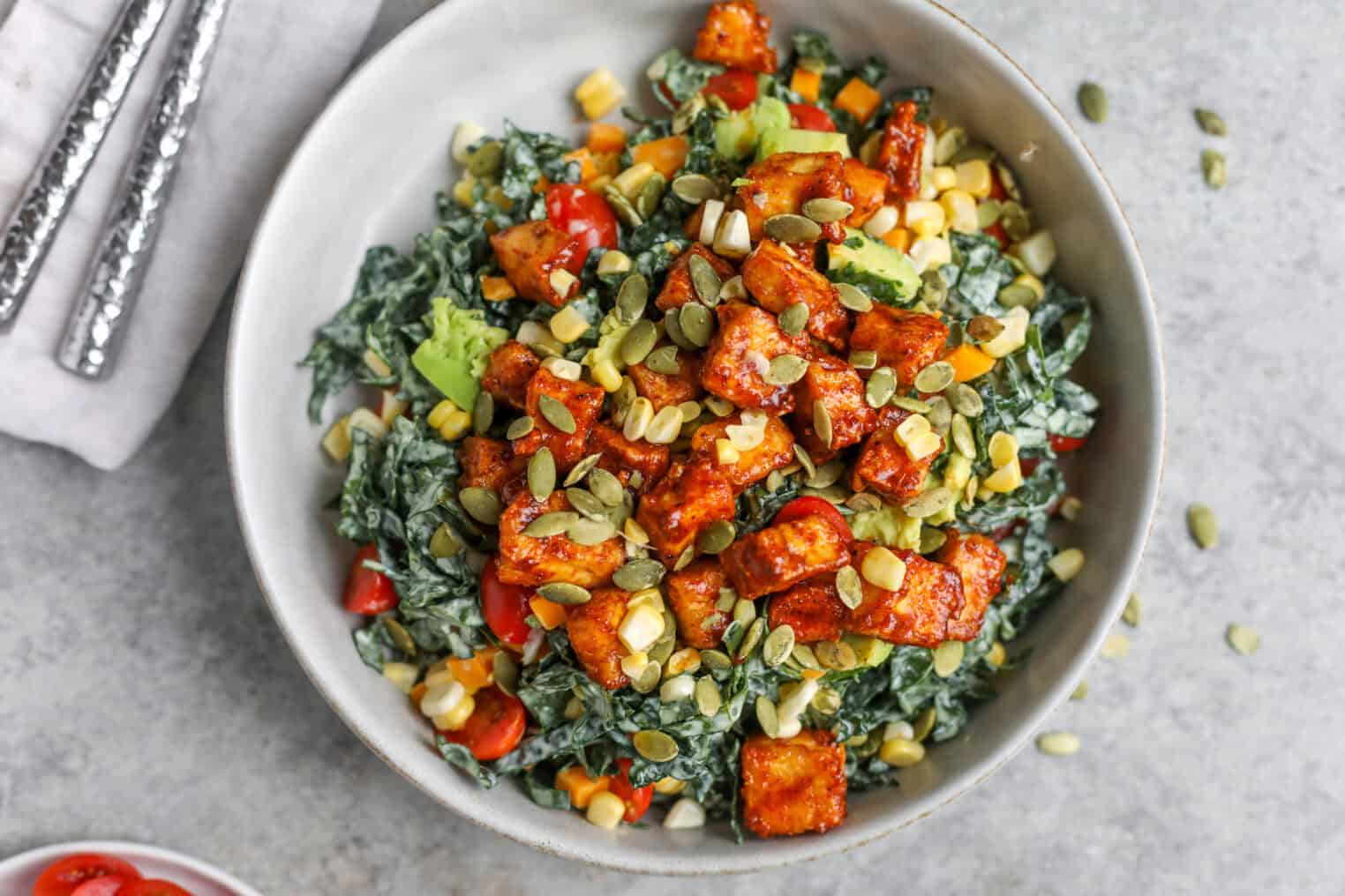 Spicy Kale Salad with Avocado Ranch 7 A Bowl Of Spicy Kale Salad With Diced Sweet Potatoes, Cherry Tomatoes, Corn, Avocado, And Pumpkin Seeds, Served On A Gray Surface With Silver Utensils Nearby.