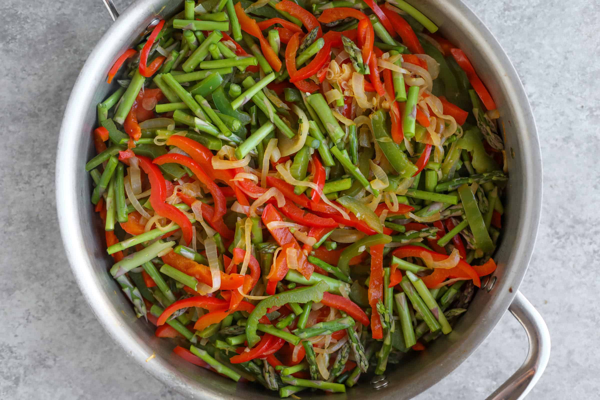 Spring Vegetable Thai Green Curry 18 A Pan Filled With A Vibrant Vegetable Medley—Sliced Red And Green Bell Peppers, Green Beans, Asparagus, And Onions—Evokes The Essence Of A Vegan Thai Green Curry. The Sautéed Vegetables Rest Enticingly On A Gray Countertop.