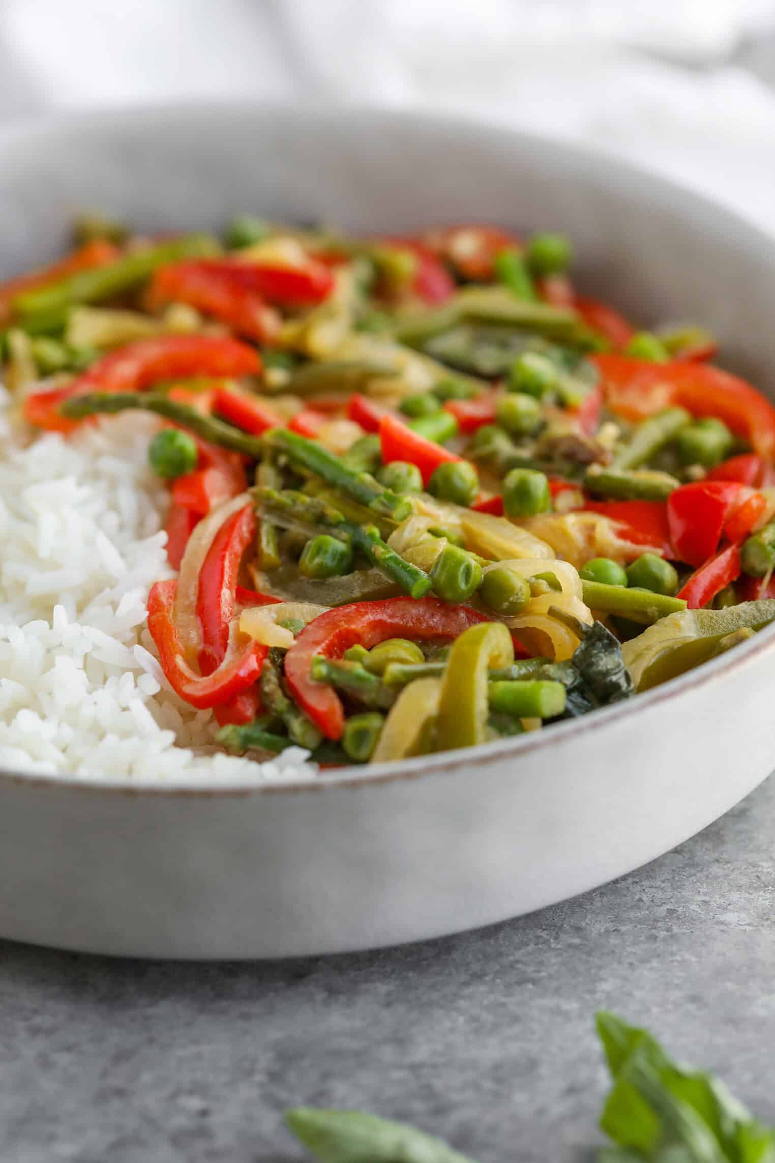 Spring Vegetable Thai Green Curry 26 A Plate Of White Rice Served With Colorful Sautéed Vegetables, Including Red Peppers, Green Beans, Peas, And Zucchini, All Gently Infused With Thai Green Curry. The Dish Is Elegantly Presented In A Round Gray Ceramic Bowl On A Gray Surface.