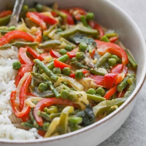 A Bowl Of Vibrant Thai Green Curry Featuring Red Bell Peppers, Peas, Green Beans, And Sliced Onions, Served Alongside Fluffy White Rice. A Spoon Rests In The Bowl On A Light Gray Surface.